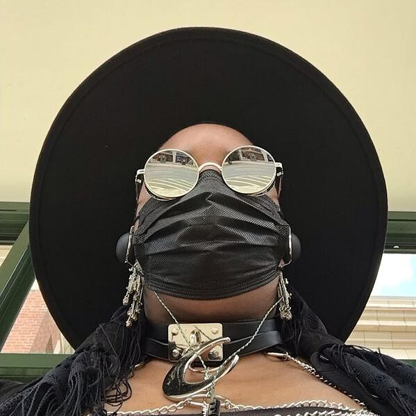 Picture of a dark-skinned person with a black mask, reflective sunglasses, a moon collar, and a wide-brimmed hat.